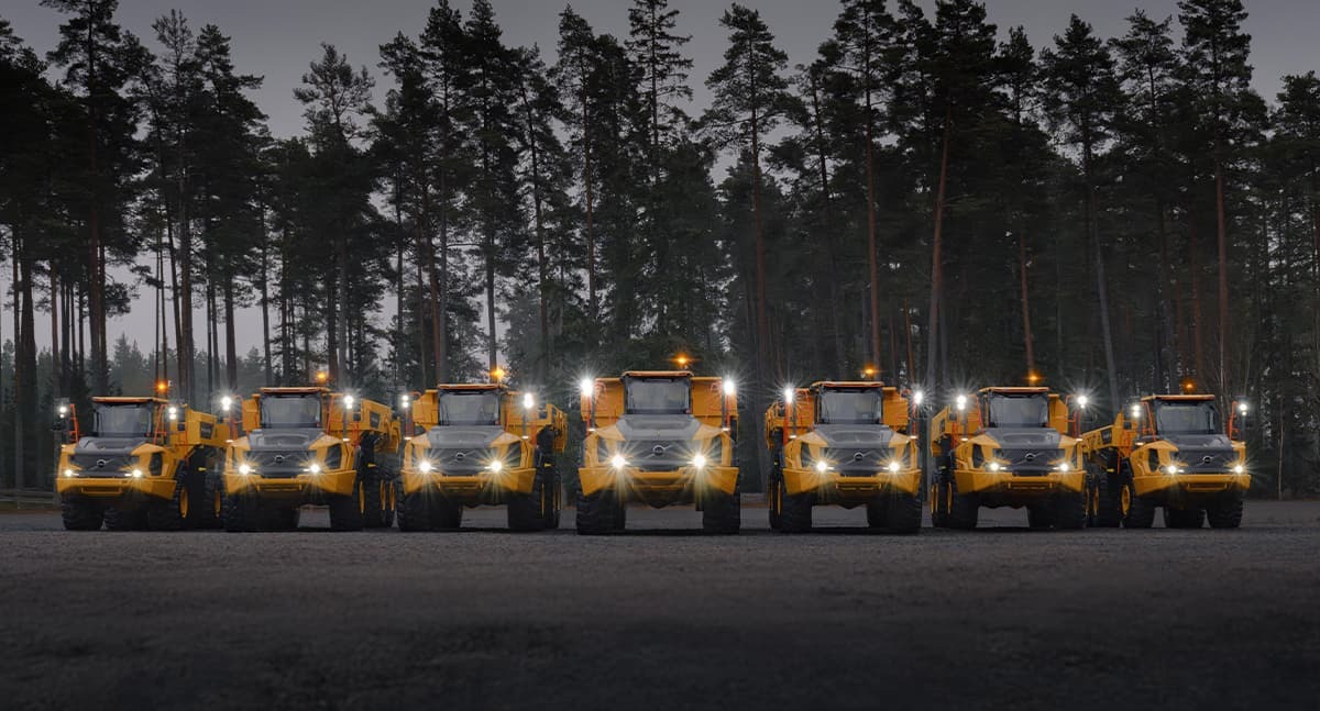 A lineup of seven Volvo articulated dump trucks at night with their headlights on.